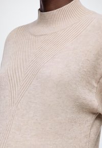 Pull en maille beige avec col montant côtelé et détail de côtes texturées en diagonale sur la poitrine et l’épaule, porté par une personne.