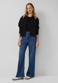 Zwarte overhemdjack met twee borstzakken, gecombineerd met brede blauwe spijkerbroeken en beige sneakers met witte accenten.