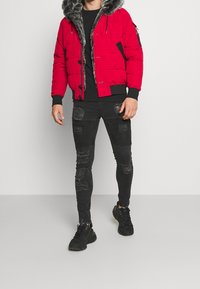 Rød polstret jakke med en pelslignende hætte, sorte detaljer på ærmerne, som er kombineret med slidte sorte skinny jeans og sorte sneakers.