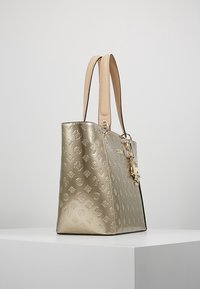 Zlatem embosovaná kožená taška přes rameno se strukturou, která má béžové uchy a dekorativní kovový prvek po straně.