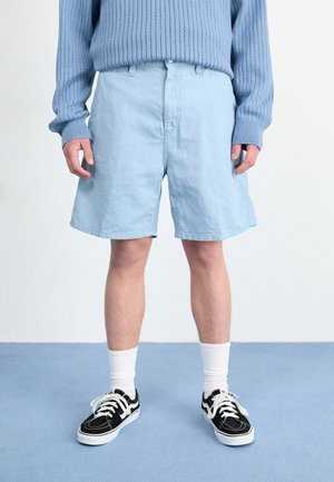 Personne portant un short bleu clair, un pull en tricot bleu, des chaussettes blanches et des baskets noires et blanches, debout sur un sol bleu.