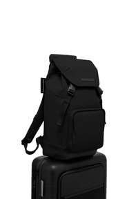 Sac à dos noir avec un rabat, fabriqué en tissu durable, comprenant une poche avant zippée et des bretelles en mesh réglables, reposant sur une valise noire.