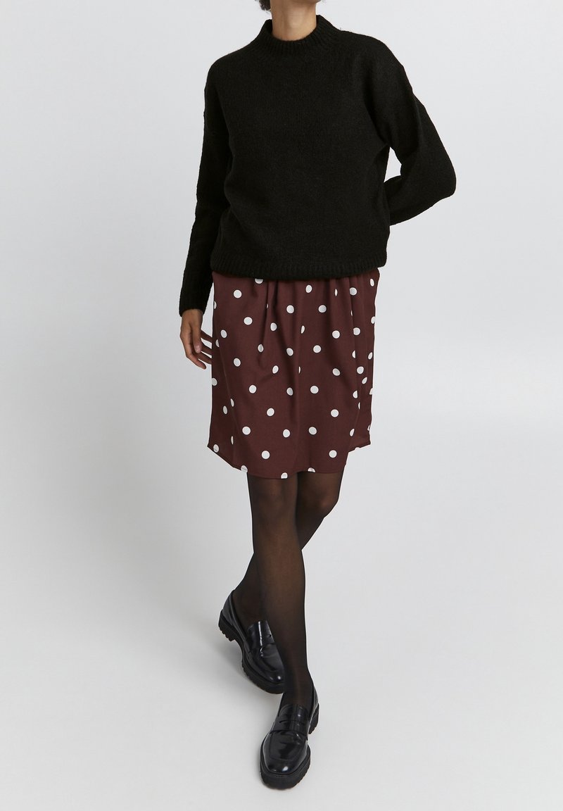 Pull en maille noir avec un col rond, associé à une jupe bordeaux à pois et des mocassins noirs, présenté sur un fond neutre.