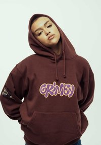 Sudadera marrón con un bolsillo frontal, que presenta un gran gráfico de "GRIMEY" en púrpura y amarillo. Tiene una capucha con cordón y un ajuste ligeramente holgado.