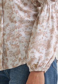 Manche de blouse de couleur claire avec imprimé floral et cheval marron, poignet élastique et détail brodé sur un jean en denim bleu.
