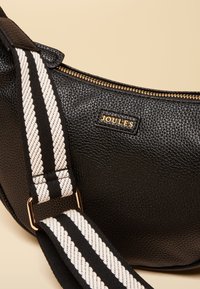 Sac en cuir noir texturé avec une bandoulière réglable rayée noir et blanc et une fermeture éclair dorée, avec un petit logo doré "Joules".