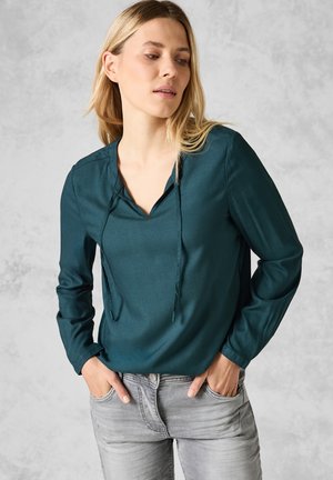 Blusa lunga teal a maniche lunghe con scollatura a V e dettaglio con coulisse. Tessuto liscio e lucido; abbinata a jeans aderenti grigio chiaro.