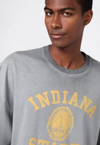 Junger Mann trägt einen grauen Pullover mit gelbem "Indiana State"-Text und einem Fort Wayne-Siegel in der Mitte auf einem weißen Hintergrund.