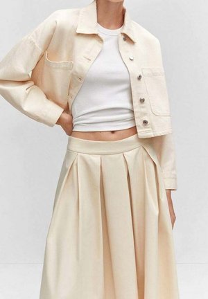 Femme portant une veste courte crème boutonnée, un haut sans manches blanc ajusté et une jupe beige plissée taille haute sur un fond uni.