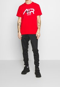 Röd bomullströja med stort vitt "AIR"-logotyp och texten "JUST DO IT.", i kombination med svarta sweatpants med vita sidodetaljer och svarta sneakers.