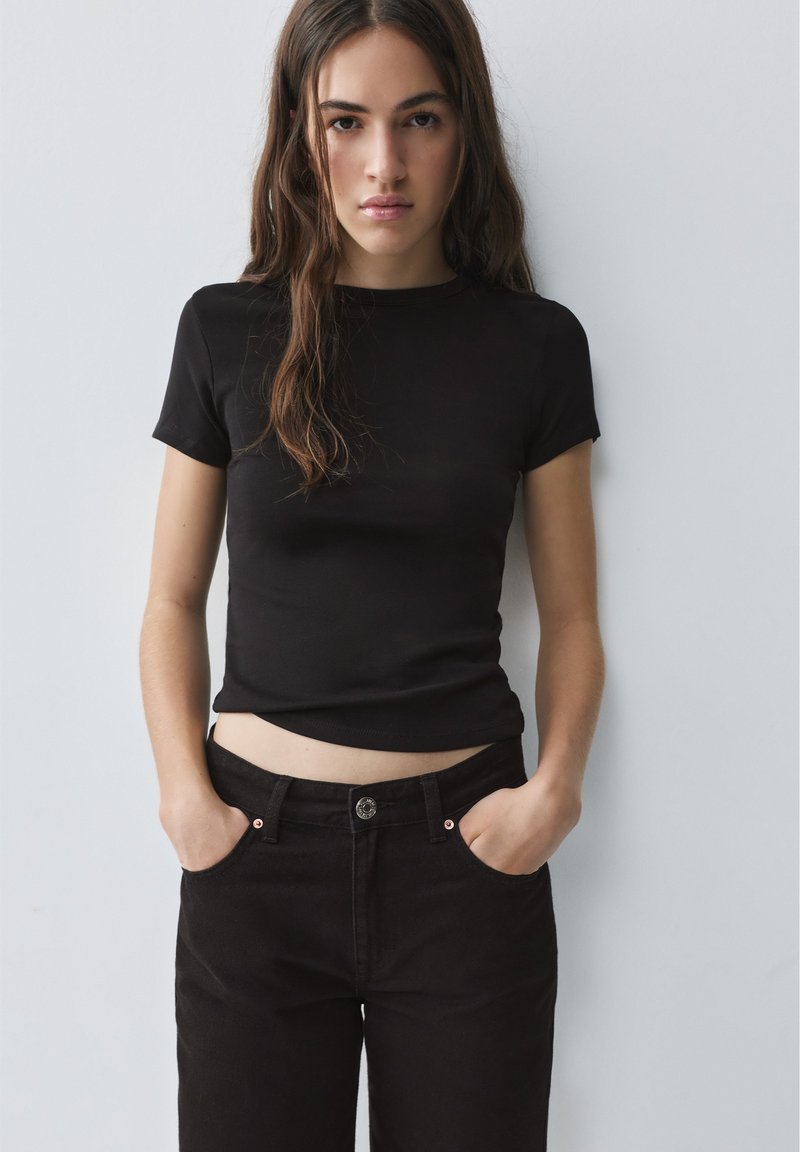 Jeune femme aux longs cheveux bruns portant un t-shirt noir ajusté et un jean noir, debout les mains dans les poches contre un mur blanc.