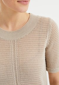 Beige kortärmad stickad topp med ribbad rund halsringning och strukturerad söm. Har ett subtilt mönster och ett lätt material.