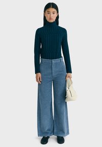 Mujer joven con cabello largo y oscuro, vistiendo un suéter de cuello alto de estampado azul oscuro, pantalones de pana de pierna ancha de color azul claro, zapatos negros, y sosteniendo un pequeño bolso beige.