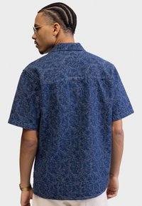 Short-sleeve blue shirt with a textured pattern, featuring a collar and a straight hem. Fabric appears lightweight and casual.