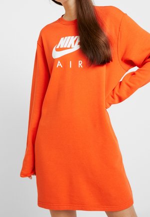 Osoba w pomarańczowej sukience Nike Air z długim rękawem i białym logo, ręka na biodrze, długie brązowe włosy na ramieniu na jednolitym tle.