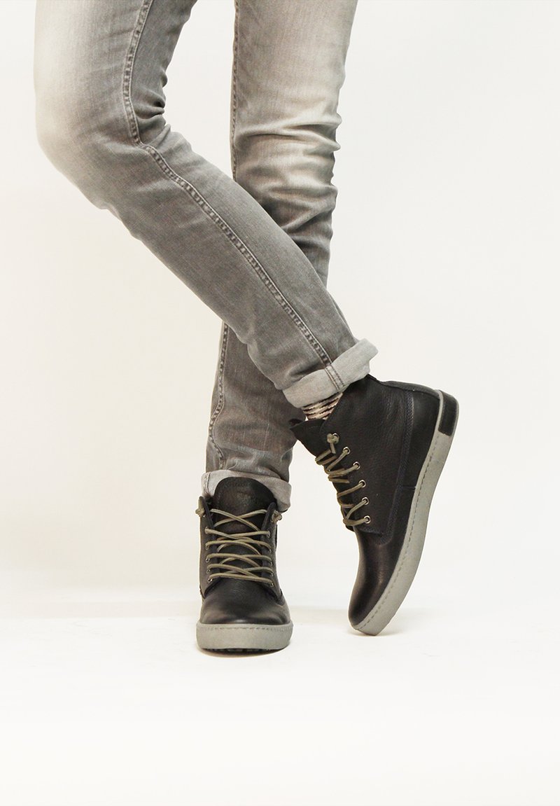 Zapatillas negras de caña alta con suelas de goma gris, que presentan cordones verdes y un exterior de cuero texturizado, combinadas con jeans grises arremangados.