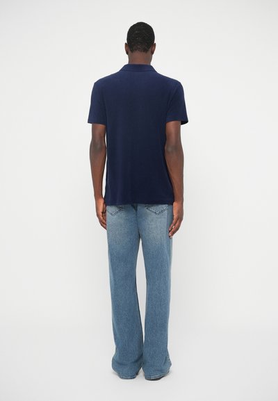 Tumši zilas krāsas polo krekls ar īsām piedurknēm, ar apkakles detalizāciju, apvienots ar gaiši zilajiem platumiem džinsiem, kurus raksturo neliela izbalēšana un vidējas augstuma piegriezums.