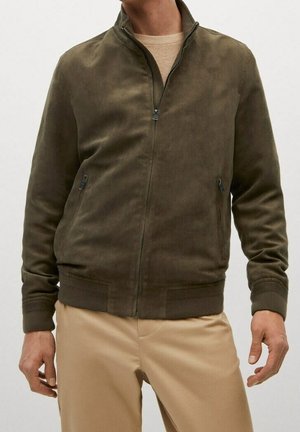 Mand iført olivengrøn ruskind jakke med lynlås, med ribstrikkede manchetter og kant, kombineret med beige bukser og beige sweater under.