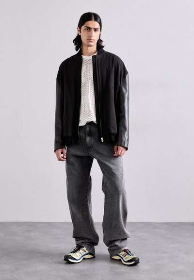 Jachetă bomber neagră cu mâneci din piele, cămașă din plasă albă, blugi gri largi și adidași negri cu galben. Design minimalist.