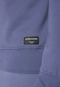 Detailný záber na fialovú látku mikiny s čiernou nášivkou označenou "BJÖRN BORG STOCKHOLM 1956" blízko švu pásu.