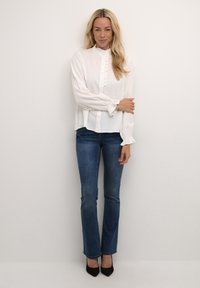 Weiße Bluse mit langen Ärmeln, rüschigem Kragen und Knopfleiste, kombiniert mit blauen Schlagjeans und schwarzen spitzen Absatzschuhen.