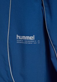 Blå sportjacka i strukturerad tyg, med Hummel-logotyp och reflekterande detaljer längs sömmarna för ökad synlighet.