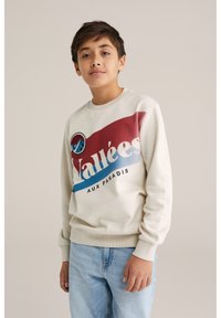 Krämfärgad sweatshirt med maroon och blå grafik "Vallées" som visar en bergslogotyp. Ribbad mudd och fåll, kombinerad med ljusblå jeans.