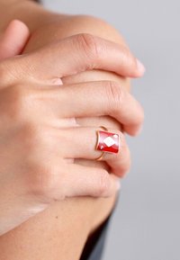 Červený smaltovaný prsteň so zlatými akcentmi, ktorý má diamantový vzor. Prsteň je hladký a dizajn je geometrický a lesklý.