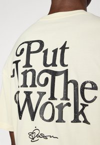 Kreemist puuvillast T-särk, millel on suur must tekst "Pane Tööle" kulunud efektiga ja allservas allkiri graafika.