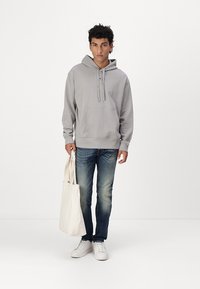 Grau Baumwoll-Hoodie mit einer Kängurutasche, kombiniert mit blauen Jeans. Das Model hält eine cremefarbene Tragetasche und trägt weiße Sneakers.