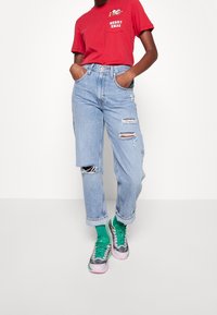 Camiseta corta roja con un gráfico de "MERRY XMAS", combinada con unos jeans desgastados de color azul claro y unas zapatillas multicolores con acentos verdes.