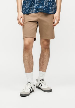 Homme portant un short beige, des chaussettes blanches, des baskets noires et blanches, et une chemise bleue à motifs, se tenant devant un fond blanc.