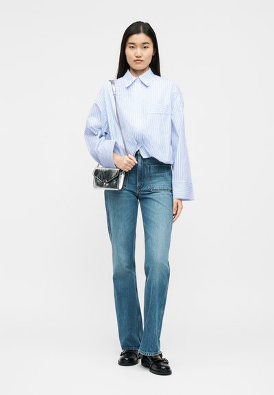 Γυναίκα που φοράει ανοιχτό μπλε ριγωτό φαρδύ πουκάμισο, μπλε τζιν παντελόνι ίσιου ποδιού, μαύρα μοκασίνια και κρατάει ασημένια τσάντα ώμου.