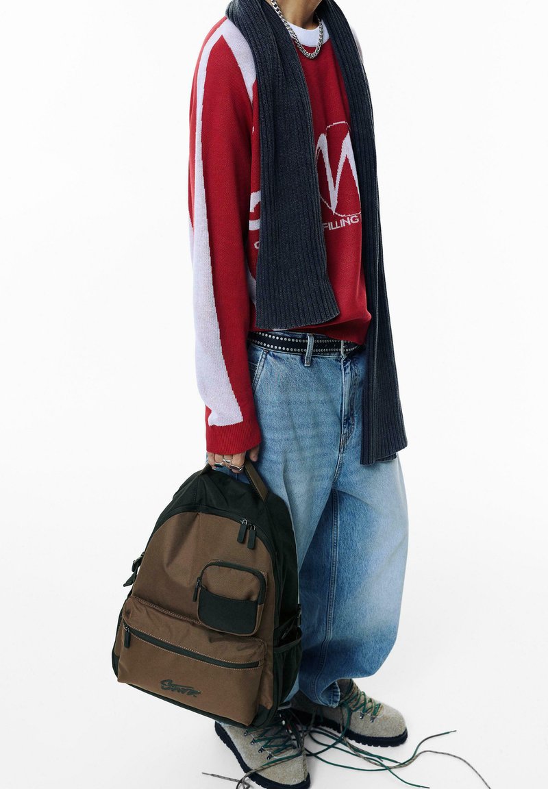 Zaino marrone e verde con molte tasche con zip, texture in tessuto, design curvo e maniglia superiore. Indossato con un maglione rosso e jeans blu.