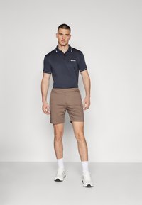 Mornarska polo majica s bijelim detaljima na ovratniku i logotipom, uparena s smeškastim šortsevima. Model nosi bijele tenisice i čarape do gležnjeva, stoji na jednostavnoj pozadini.