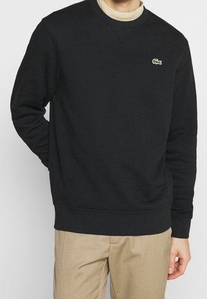 Mann trägt schwarzen Sweatshirt mit kleinem grünem Krokodil-Logo auf der Brust, beige Hose und hellen Rollkragen darunter.