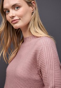 Młoda kobieta z blond włosami, ubrana w prążkowany sweter w kolorze przydymionego różu oraz małe srebrne kolczyki w kształcie kół, patrzy w stronę kamery na tle ciemnego tła.