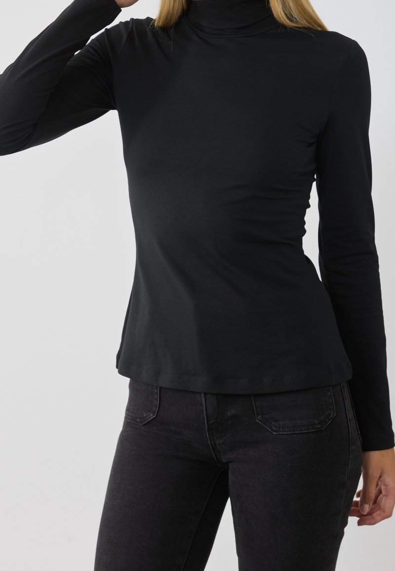 Schwarzes langärmliges Rollkragenshirt in einer figurbetonten Passform aus weichem Stoff. Nahtloses Design und glatte Textur. Kombiniert mit dunklen Jeans.