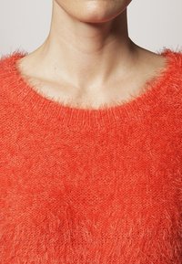 Röd, fluffig tröja med en bred, rund halsringning. Mjuk textur, lösa fibrer och ribbade ärmslut ger ett mysigt intryck.