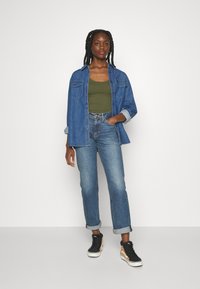Denim shirt in blauw, groene tanktop, high-waisted blauwe spijkerbroek en zwarte high-top sneakers met oranje accenten. Model staat tegen een witte achtergrond.