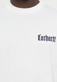 White cotton T-shirt featuring a small dark blue "Carhartt" logo embroidered on the left chest area. Round neckline, short sleeves.