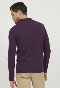 Polo à manches longues en coton violet profond, featuring un col classique et une coupe décontractée avec des fentes latérales pour plus de liberté de mouvement.