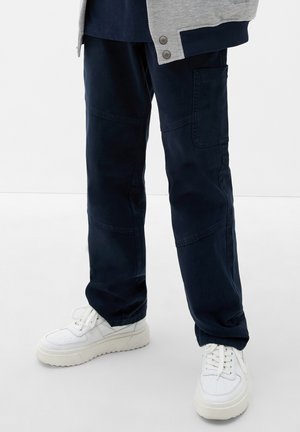 Pantalon cargo bleu marine avec poches latérales, fabriqué en matière durable. Associé à des baskets blanches avec une semelle épaisse.