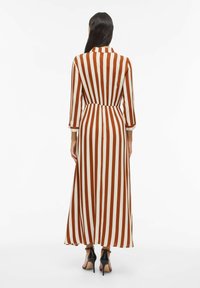 Robe longue rayée en marron et crème avec une taille cintrée, une patte de boutonnage et des manches longues ; associée à des talons noirs.