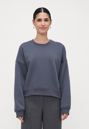 Een vrouw met donker haar in een lage knot draagt een losse donkerblauwe sweatshirt en grijze pinstripe broek op een witte achtergrond, met het gezicht naar voren.