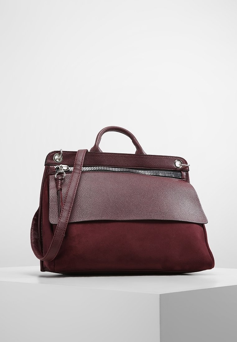 Sac à main en cuir et daim bordeaux foncé avec poignée supérieure, rabat avant, poche zippée et bandoulière détachable sur un piédestal blanc.