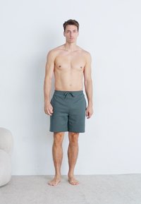 Grå drawstring svømmebukser med bløt tekstur, med en avslappet passform og sidelommer, båret av en stående hannmodell mot en ensfarget bakgrunn.