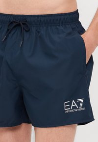 Námorné plavky s elastickým pásom, nastaviteľnou šnúrkou a bočnými vreckami. Obsahujú potlačené logo EA7 v striebornej farbe.