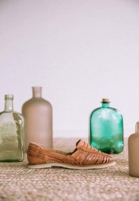 Zapato de cuero trenzado marrón con suela plana, que presenta un diseño abierto en la parte trasera y un patrón intrincado, colocado cerca de varias botellas de vidrio sobre una superficie texturizada.