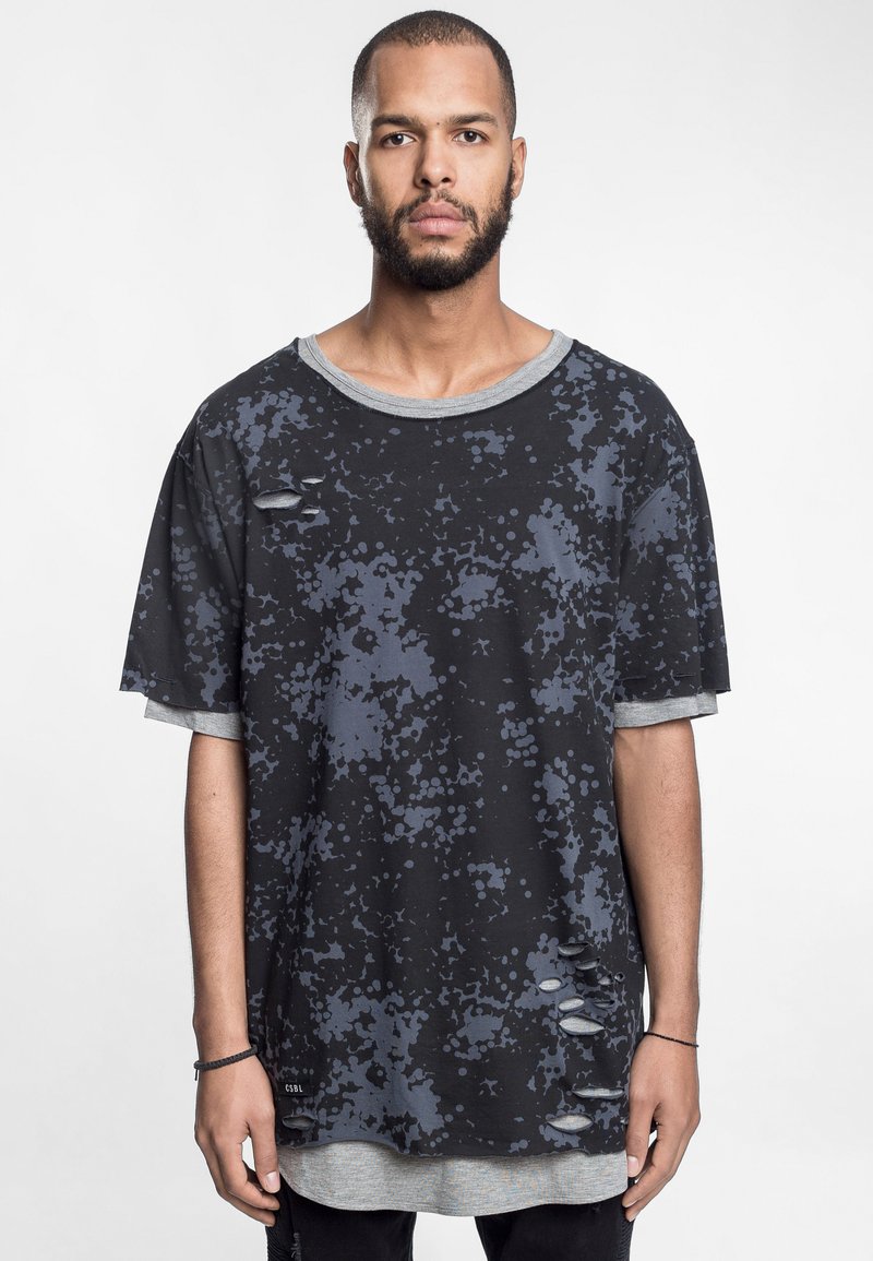 Sort oversized t-shirt med grå spraglet mønster, ribbet halsudskæring og slidte detaljer på forsiden. Laget over en grå langærmet skjorte.
